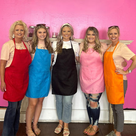 Five smiling women wearing red, blue, black, pink and orange aprons posing together against a bright pink studio wall in a local craft/art workshop