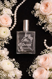 Bridal flat-lay of a clear engraved perfume bottle with floral script and a wedding date, framed by pale pink roses, white ranunculus, baby's breath and pearl necklaces on a black background.