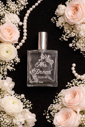 Bridal flat-lay of a clear engraved perfume bottle with floral script and a wedding date, framed by pale pink roses, white ranunculus, baby's breath and pearl necklaces on a black background.