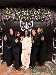 Five women smiling in front of a purple-and-white floral wall with a decorative butterfly logo; center woman in white wearing a veil holds a perfume bottle while her friends in black also display bottles — indoor bridal shower bachelorette celebration.