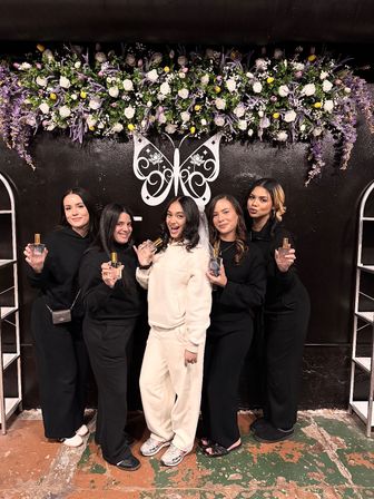 Five women smiling in front of a purple-and-white floral wall with a decorative butterfly logo; center woman in white wearing a veil holds a perfume bottle while her friends in black also display bottles — indoor bridal shower bachelorette celebration.