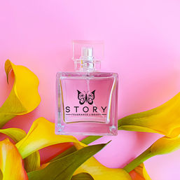 Clear glass perfume bottle with a butterfly emblem on a pastel pink background, flat-lay surrounded by bright yellow calla lilies — spring floral fragrance still life.