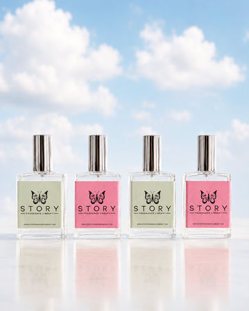 Four clear fragrance bottles with silver caps in a row on a glossy marble surface, pastel pink and mint labels with a butterfly logo, set against a bright blue sky with fluffy white clouds.