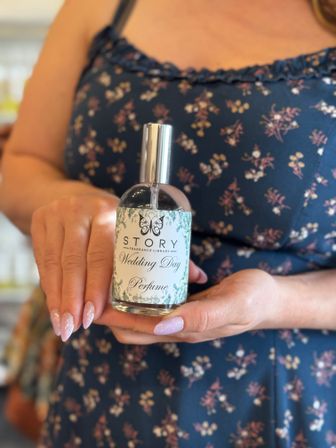 Close-up of hands with glittery pink almond nails holding a Wedding Day perfume bottle with a floral label against a navy floral dress, bridal fragrance