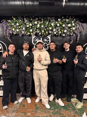Six men in casual streetwear posing indoors at a boutique-style black wall, each holding a perfume bottle beneath a large purple and white floral garland and decorative butterfly wall art.