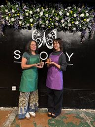 Two people in green and purple aprons smiling and holding glass perfume bottles beneath a cascading white, purple and yellow floral garland on a black wall with a decorative butterfly mural — indoor boutique perfume and floral studio scene.