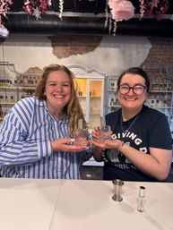 Two smiling people at a perfume-making workshop counter holding matching fragrance bottles, with hanging floral decor, exposed brick wall and shelves of scent bottles in a cozy fragrance studio interior.