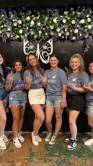Six women in matching blue graphic shirts and casual shorts or skirts stand side-by-side indoors, smiling and holding perfume bottles in front of a black wall with a white butterfly logo and a lush purple, white and yellow floral garland — casual group photo with floral backdrop.