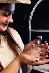 Close-up of a smiling woman in a hat holding a personalized engraved glass perfume bottle with floral etching and date, against a dark backdrop and metal shelving — elegant fragrance detail shot.