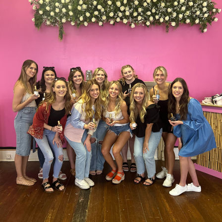 Group of eleven women smiling and posing indoors holding small perfume bottles in front of a bright pink wall with a white rose floral backdrop — fun girls’ day photo