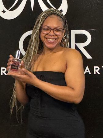 Smiling woman with long blonde braids and glasses in a strapless black dress holding a perfume bottle in front of a black logo backdrop — beauty product portrait.