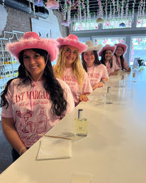 Five women wearing pink fluffy cowboy hats and matching pink "Last Margarita as a Señorita" shirts smile along a white counter in a bright, flower-decorated scent studio, each with a small perfume bottle — playful bachelorette celebration.