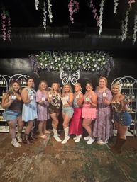 Group of women smiling and holding perfume bottles inside a floral-decor boutique fragrance bar with hanging wisteria and decorative butterfly wall art