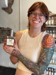 Smiling person with short red hair and round glasses holding a clear fragrance bottle inside a boutique, showing a colorful full-sleeve tattoo of a ship, swirling sun and floral designs.