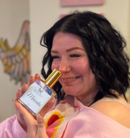 Smiling woman with black curly hair and a nose ring holding a fragrance bottle with a gold cap close to her face in a cozy indoor setting with colorful wall art