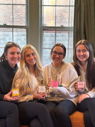 Four smiling friends seated on a couch by tall paned windows, each holding a decorative perfume bottle with gold caps in a cozy, sunlit indoor setting.
