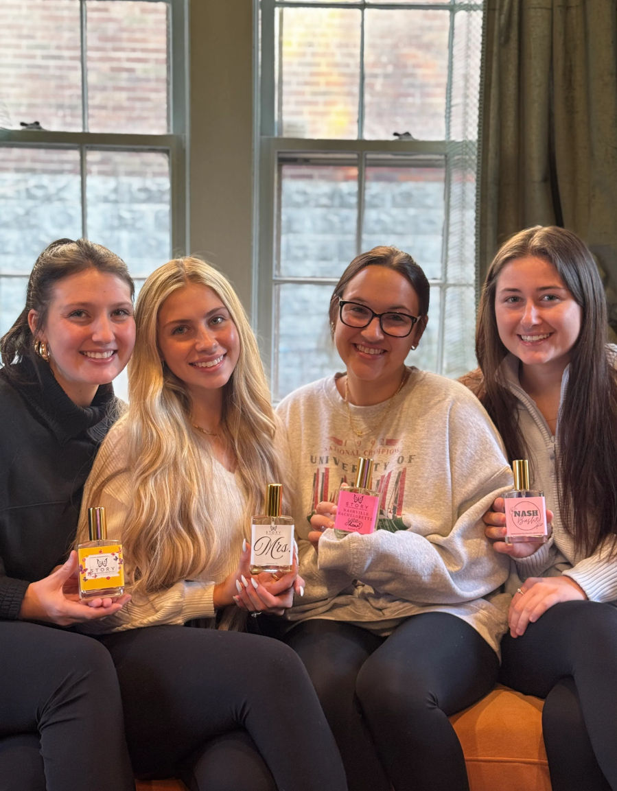 Four friends sitting on a couch by tall windows, smiling and holding colorful perfume bottles in a cozy indoor setting.