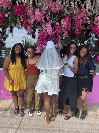 Group photo of five women in front of a vibrant pink floral storefront wall; bride-to-be in a white cowboy hat and veil faces away while friends in casual summer outfits smile and pose, one holding a small bottle — playful bachelorette moment.