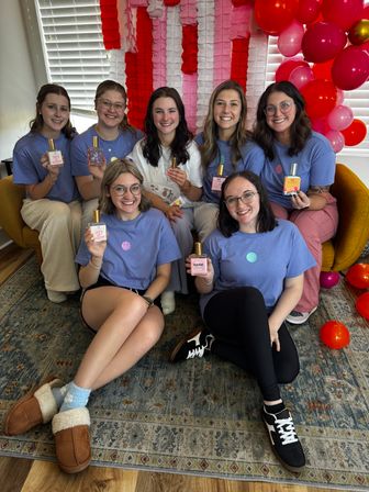 Seven friends in matching lavender shirts pose in a cozy living-room perfume party, each holding a fragrance bottle in front of a pink-and-red balloon and paper-streamer backdrop.