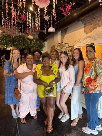 Group photo of eight smiling women in a flower-adorned event space with hanging pink blossoms, exposed brick and black ductwork; woman in the center wears a bright yellow ruffle dress and holds a leopard-print clutch.
