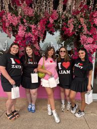 Five friends smiling on a city sidewalk in front of a dense pink floral storefront display, wearing playful "I ♥" band-name T-shirts (Zayn, Louis, Hunter, Liam, Harry) and holding white gift bags.