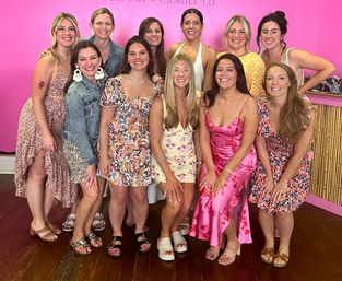 Twelve smiling women in colorful summer dresses and sandals pose together on a wooden floor in front of a bright pink boutique wall and bamboo counter, cheerful friends celebrating indoors.
