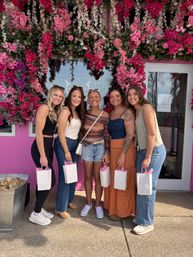 Five friends posing outside a pink storefront with a cascading pink-and-white floral wall backdrop, each holding a small white gift bag — outdoor group photo.