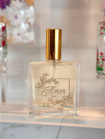 Clear square glass perfume bottle with shiny gold cap labeled "Amor" with a hand-drawn botanical wreath and small date "10.18.25", displayed on a white shelf — elegant boutique fragrance.