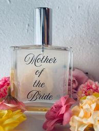 Glass perfume bottle labeled 'Mother of the Bride' with a shiny silver cap, surrounded by pink, yellow and peach flowers on a white background, wedding-themed decorative shot.