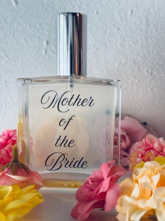 Clear perfume bottle labeled 'Mother of the Bride' with a silver cap nestled among pastel pink, yellow and peach flowers — a bridal gift and wedding-day accessory.