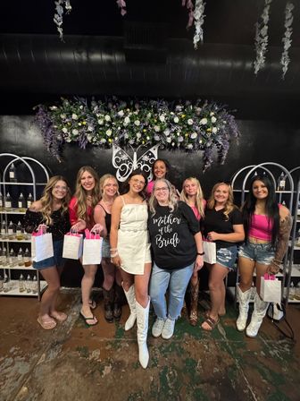 Bachelorette party group photo indoors — bride in a white dress and cowboy boots with friends holding gift bags in front of a purple-and-white floral wall and butterfly decor.