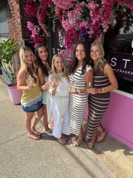 Five smiling women holding perfume bottles on a sunny sidewalk in front of a pink boutique storefront draped with cascading pink bougainvillea, cheerful summer fragrance-shopping scene.