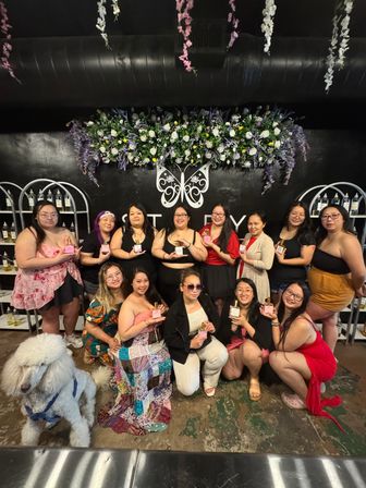 Group of women posing at an indoor perfume-making workshop in front of a decorative butterfly and floral wall, each holding a pink fragrance bottle, with a large white dog seated at the front.