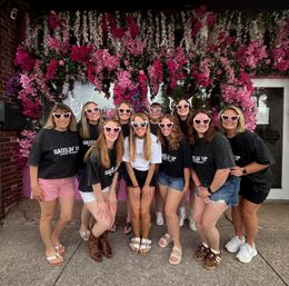 Group of ten women in matching dark tees and heart-shaped pink sunglasses smiling for a photo in front of a vibrant pink-and-white flower wall at a storefront.