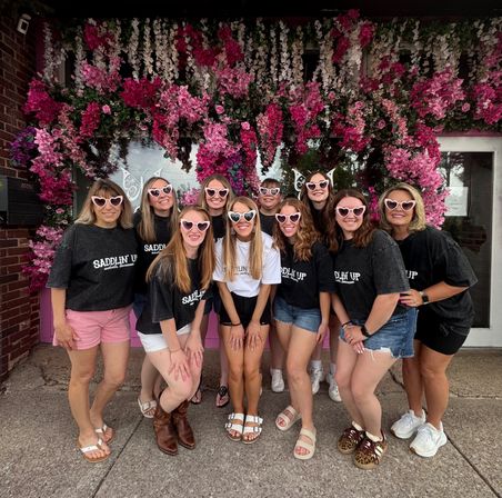 Group of ten women in matching dark tees and heart-shaped pink sunglasses smiling for a photo in front of a vibrant pink-and-white flower wall at a storefront.