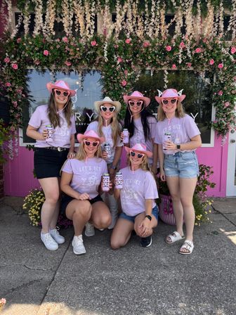 Six women in matching lavender 'Let's Get Rowdy' shirts wearing pink cowboy hats and heart-shaped sunglasses, holding canned drinks and posing in front of a pink storefront framed by hanging white and pink flower garlands.