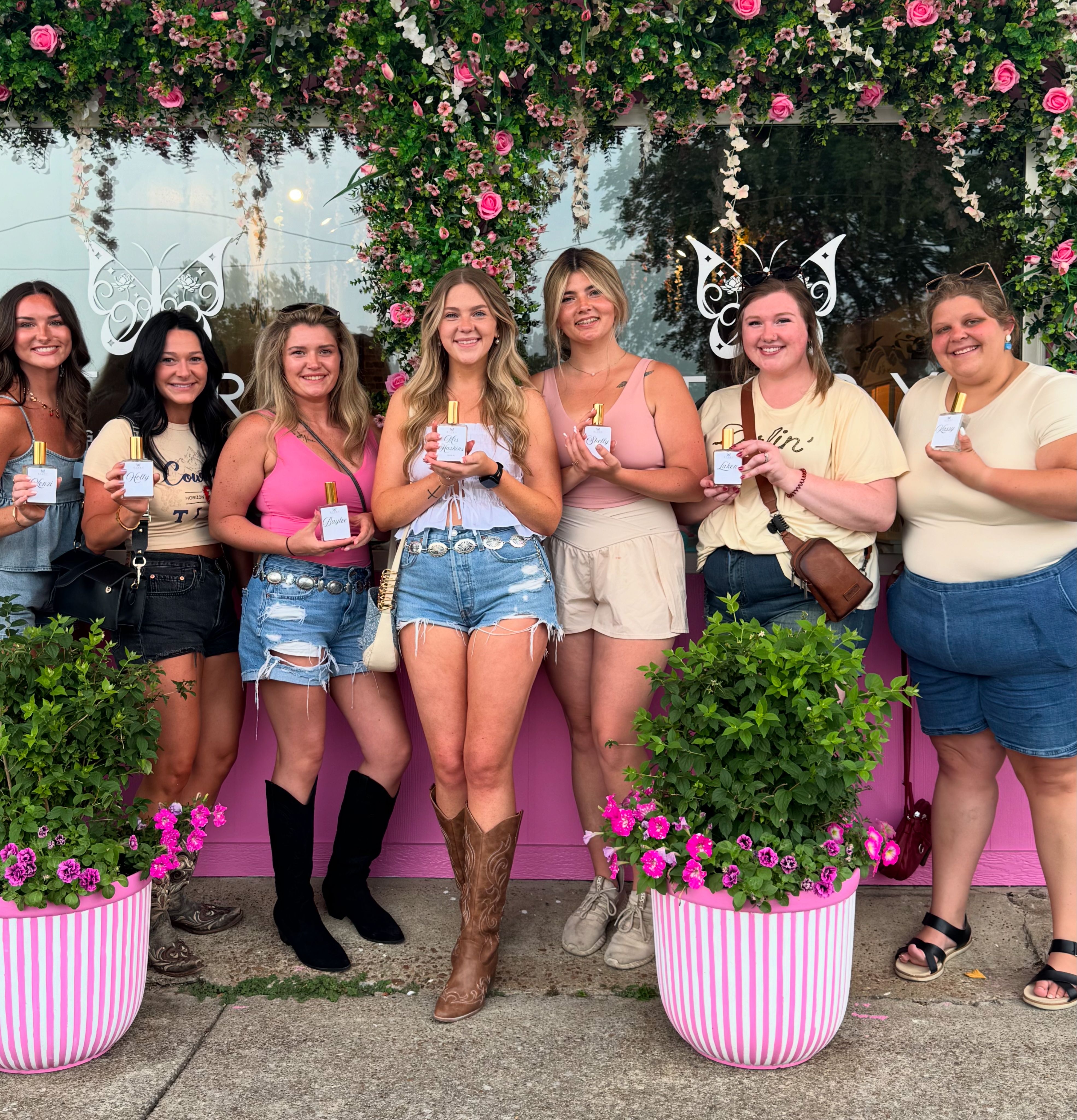 Seven women smiling and posing outside a pink, flower-adorned storefront holding perfume bottles; summer denim shorts, boots and bright floral backdrop