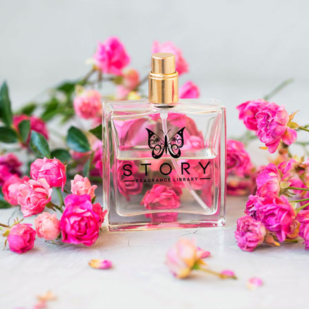 Clear square perfume bottle with a gold sprayer and butterfly motif, surrounded by vibrant pink rose blossoms on a light tabletop, floral fragrance flatlay
