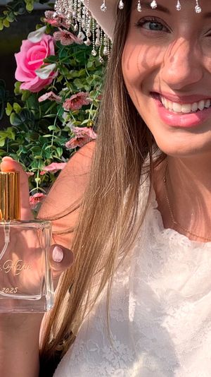 Sunlit close-up of a smiling woman in white lace holding a glass perfume bottle with a gold spray cap, beaded hat shadow on her face and pink floral backdrop