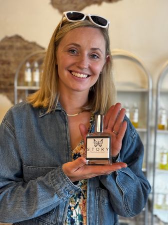 Smiling woman in a denim jacket with sunglasses on her head holding a clear perfume bottle with a decorative label inside a boutique fragrance shop with display shelves in the background