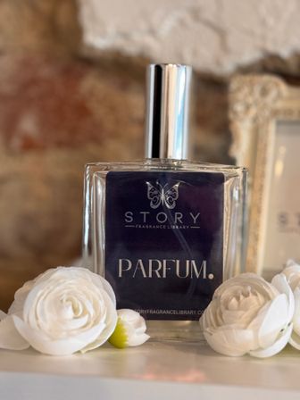 Glass perfume bottle with a shiny silver cap on a shelf, flanked by white ranunculus-style flowers and set against a blurred rustic brick background.