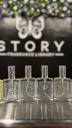 Cheeky row of clear perfume spray bottles lined up on a shiny counter at a boutique fragrance display, with a blurred black feature wall and floral-accented shelving in the background.