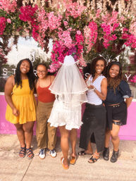Five friends smiling and posing in front of a vibrant pink floral storefront display, one wearing a white veil and cowboy hat for a bachelorette celebration, surrounded by cascading pink blooms.