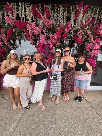 Six smiling women pose on a sunny sidewalk outside a boutique, standing under a lush pink-and-white floral arch and each holding a small glass perfume bottle — casual summer outfits, sandals and sunglasses.