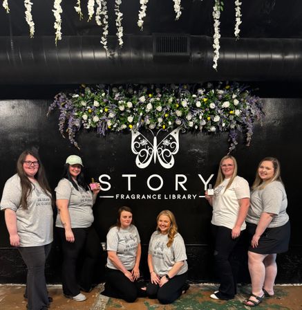 Group photo of six women in matching gray t-shirts posing indoors under hanging white wisteria and a purple-and-white floral installation in front of a black wall with a decorative butterfly logo at a fragrance shop.