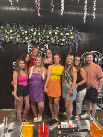 Smiling group of nine people posing under hanging floral garlands and a purple-and-white flower wall inside a DIY candle-making studio, with colorful wax jars, dye bottles, and tools on the worktable in the foreground.