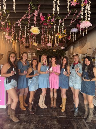 Eight smiling women in denim and pastel dresses wearing cowboy boots and holding perfume bottles inside a rustic brick boutique with hanging pink and white floral ceiling decor