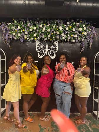 Five friends smiling and posing at an indoor boutique photo wall, each holding a perfume bottle beneath a purple-and-white floral garland and white butterfly mural.