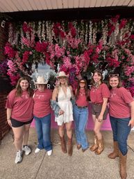 Six friends wearing cowgirl boots — five in matching pink tees and one in a white dress and cowboy hat — pose on a sidewalk in front of a storefront covered in cascading pink and white flowers.