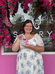 Smiling woman in blue floral dress and sash holding a perfume bottle in front of a pink storefront framed by cascading pink flowers.